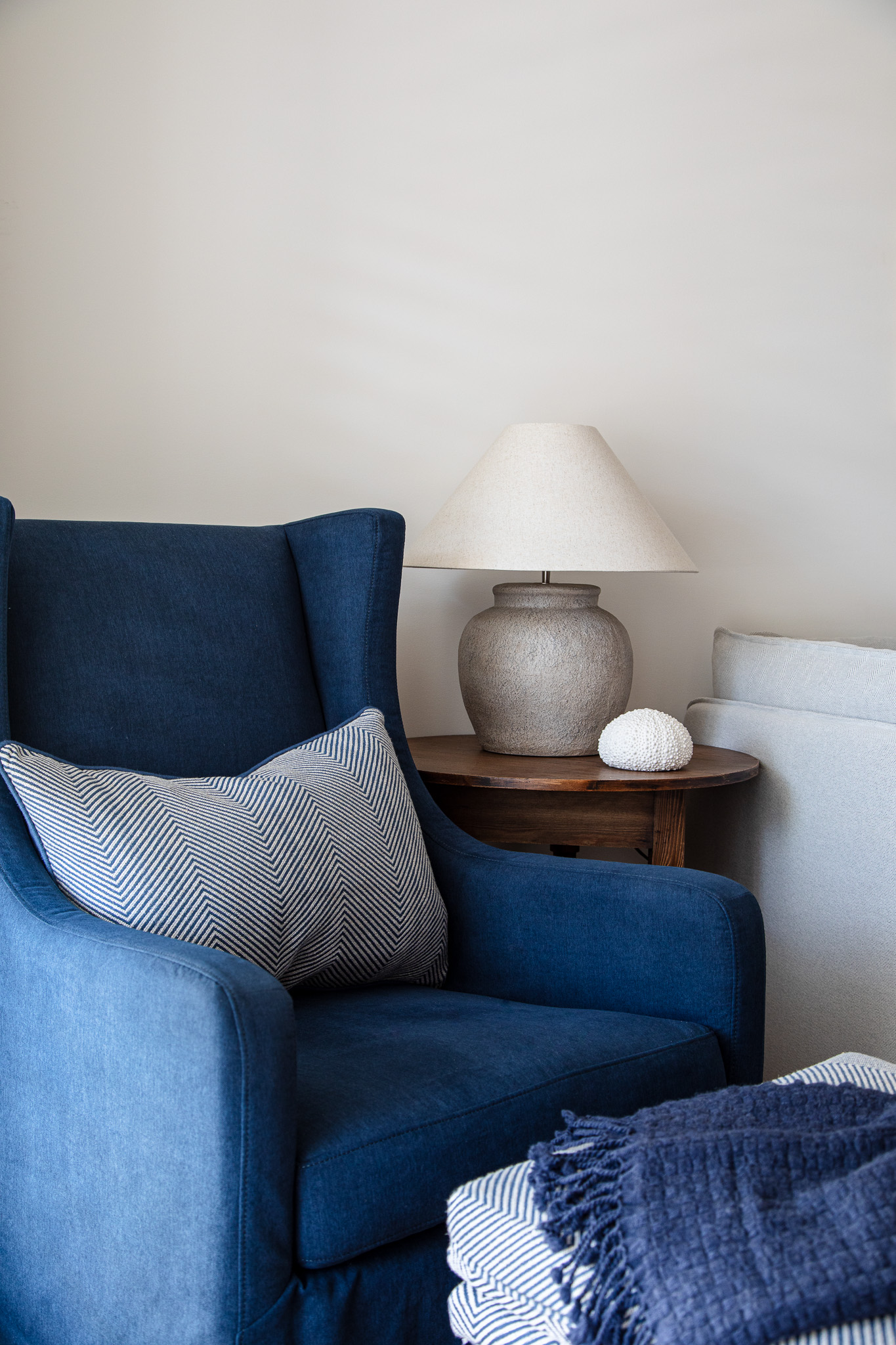 A blue armchair with striped pillows sits next to a wooden side table holding a beige lamp and a decorative white object. In the background, a gray sofa is partially visible, complementing nearby wardrobes & linen cupboards.
