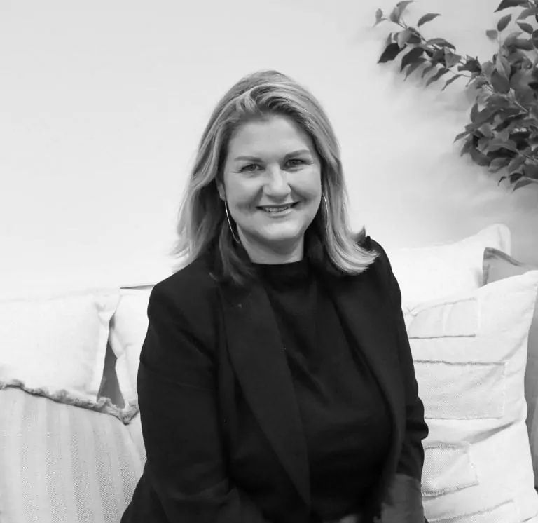 A woman, resembling Danielle Rose who founded Home Remedy, with shoulder-length hair, wearing a dark blazer and top, sits on a couch with cushions. A leafy plant is in the background. The image is in black and white.