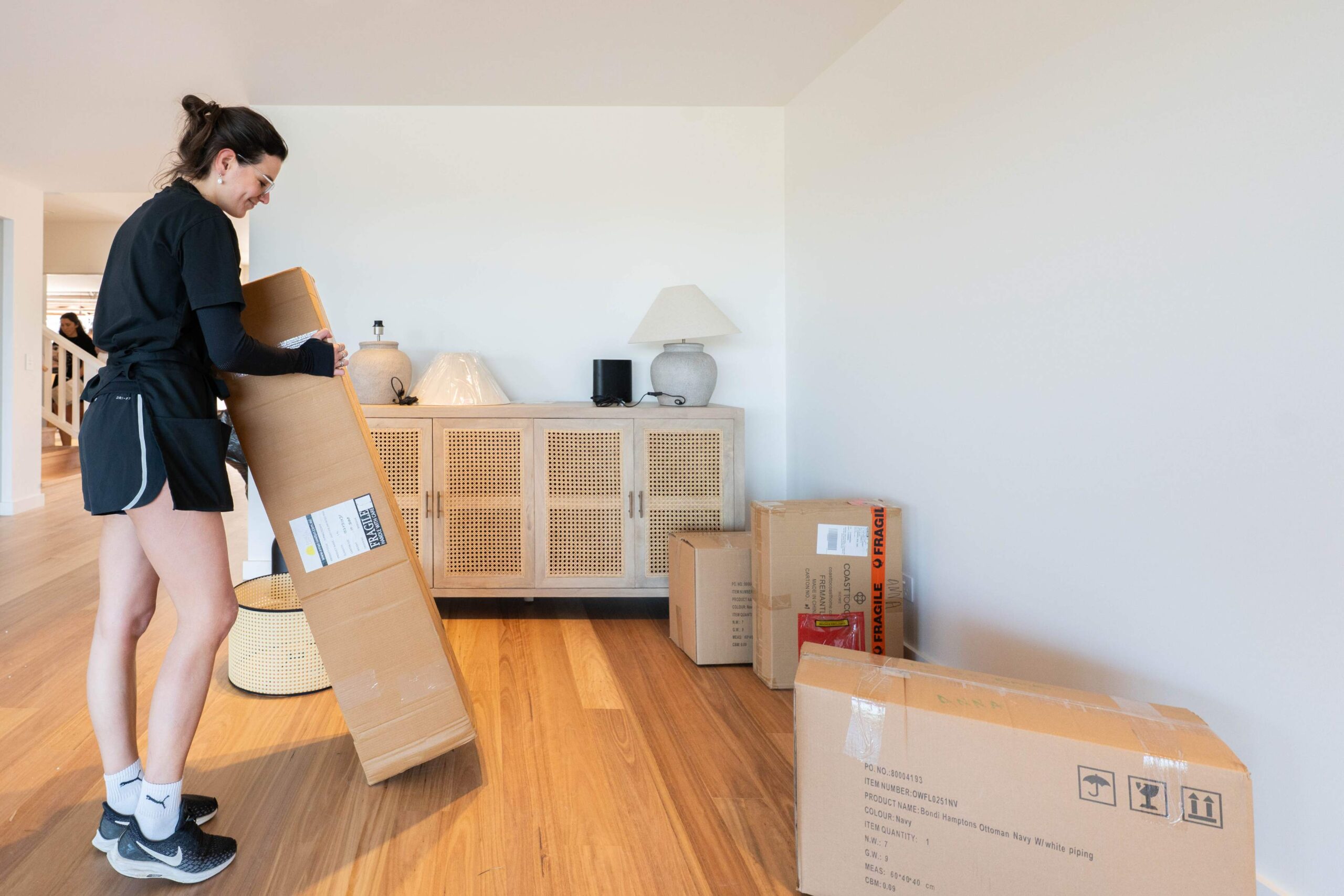 A woman in athletic wear lifts a cardboard box in a room with wooden floors, embracing decluttering and downsizing. Several other boxes are scattered around, and a sideboard with decor items is against the wall.