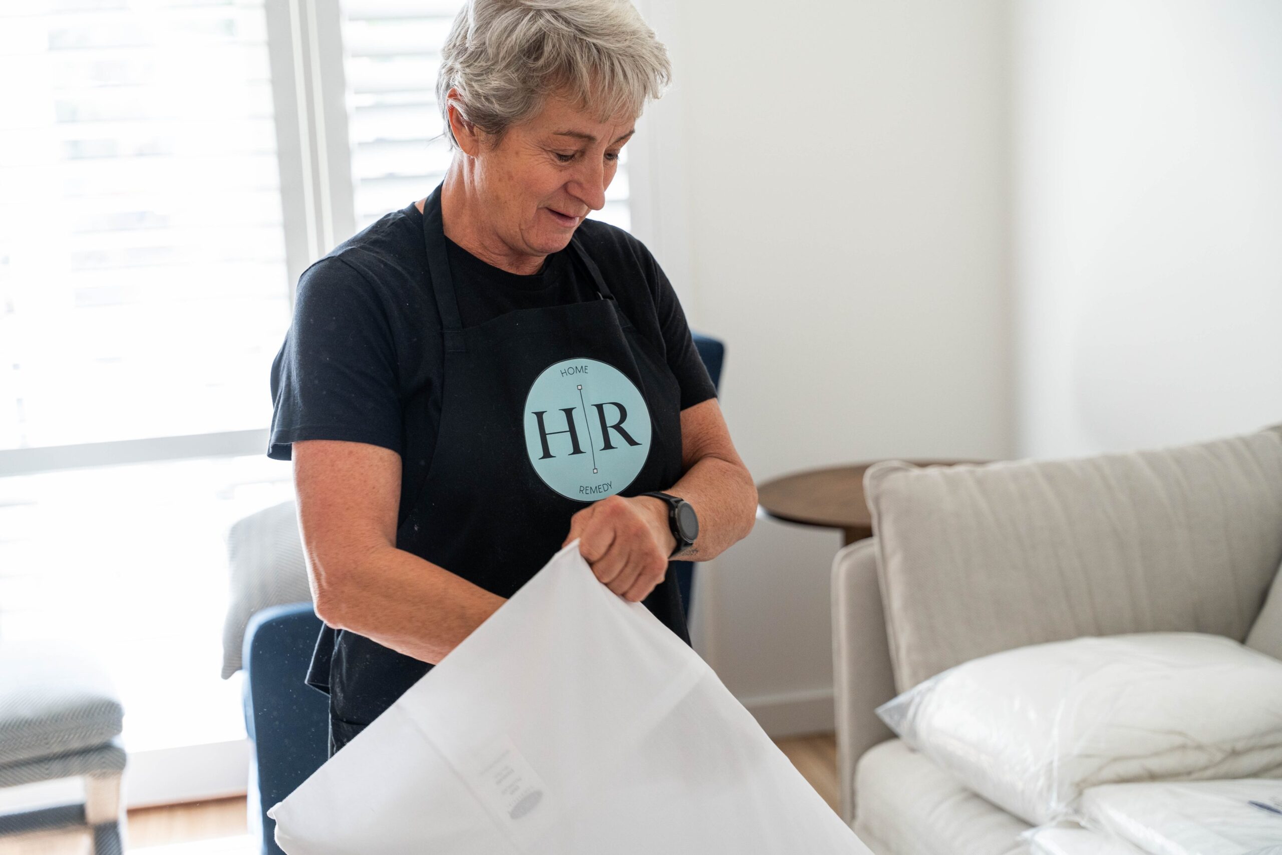 A person wearing an apron labeled "HR" smooths out a pillowcase in a bright, tidy room with a sofa and cushions, offering downsizing support in a comfortable setting.