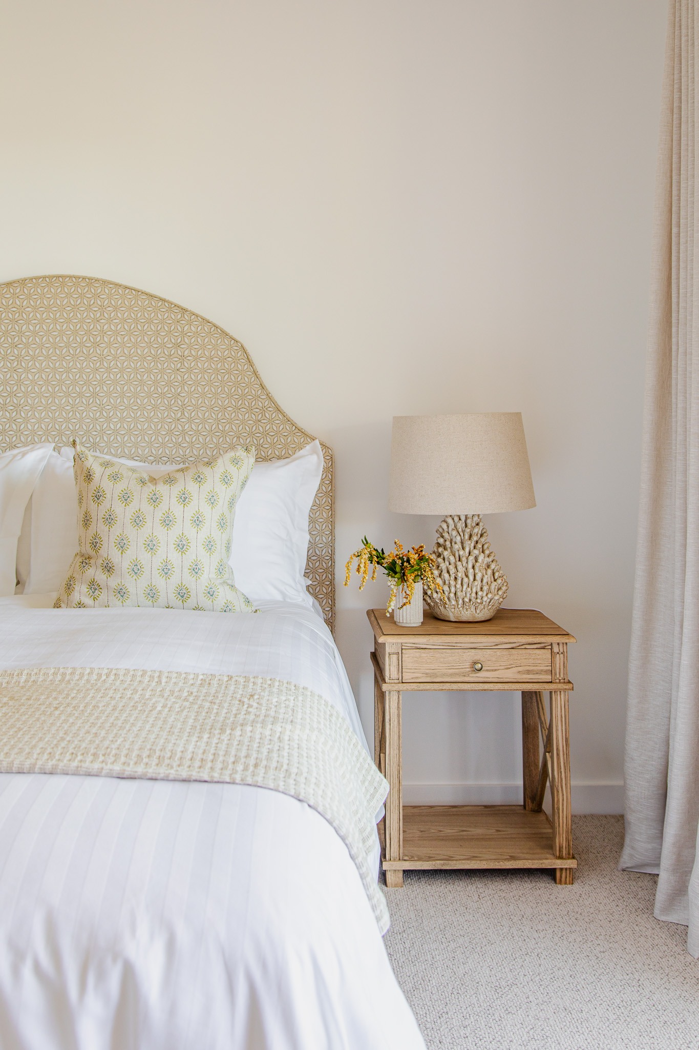 A neatly made bed with patterned pillow and blanket sits beside a wooden nightstand holding a beige lamp, a small vase with flowers, and a book—creating the perfect cozy retreat for trying any favorite home remedy in this bright, neutral-toned bedroom.