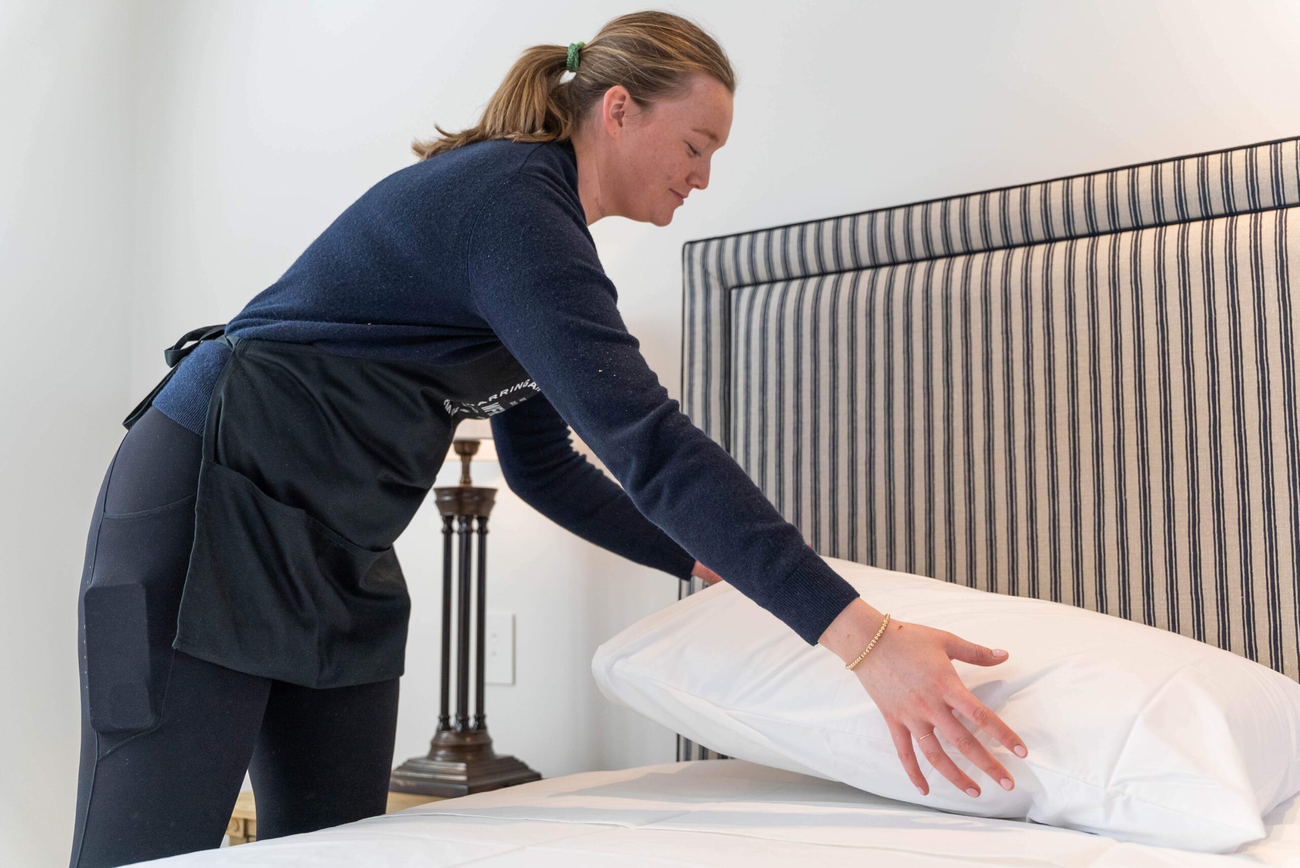 A woman in a dark sweater and apron is placing a pillow on a neatly made bed with a striped headboard, showcasing her home remedy for creating a cozy, well-lit room.