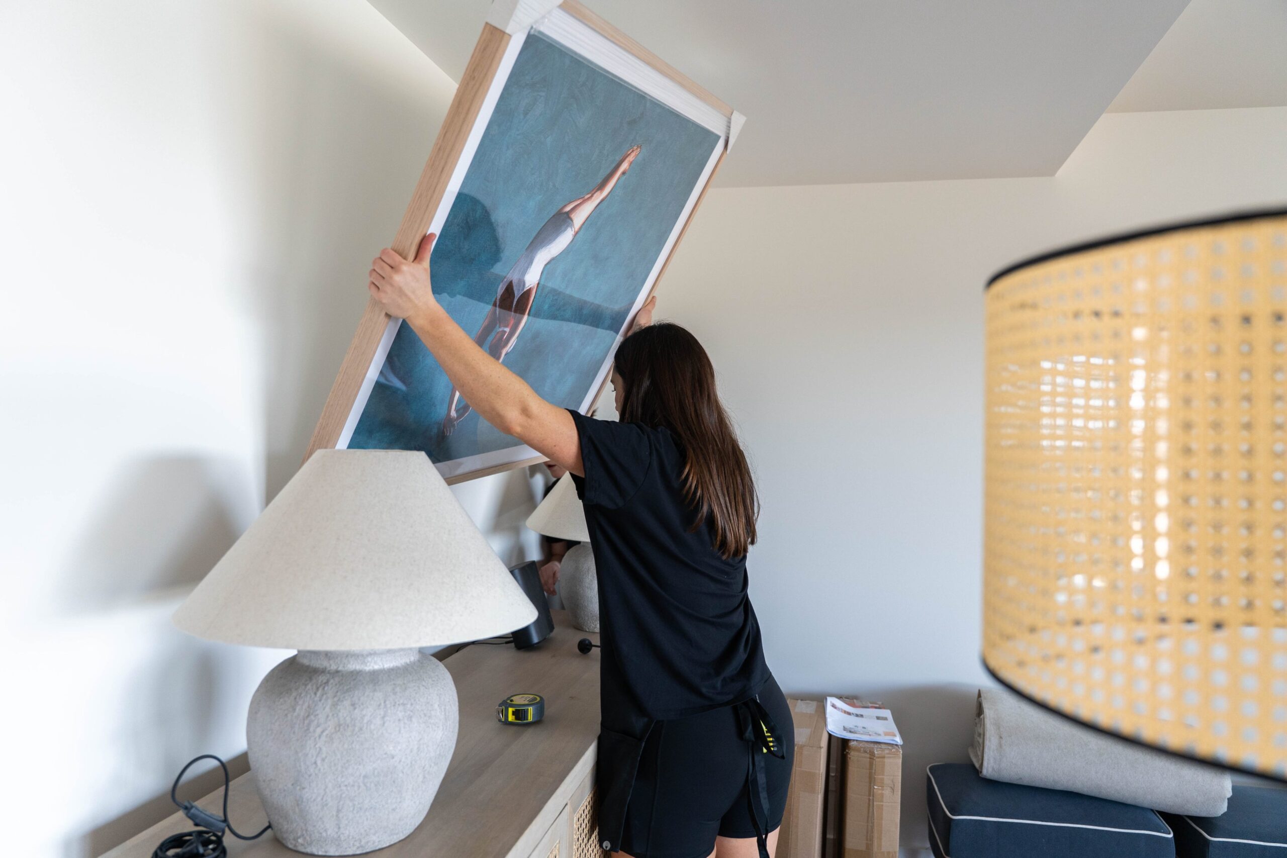 A person in black clothes hangs a framed artwork above a beige table with a lamp, tape measure, and boxes nearby a simple home remedy to refresh any space.