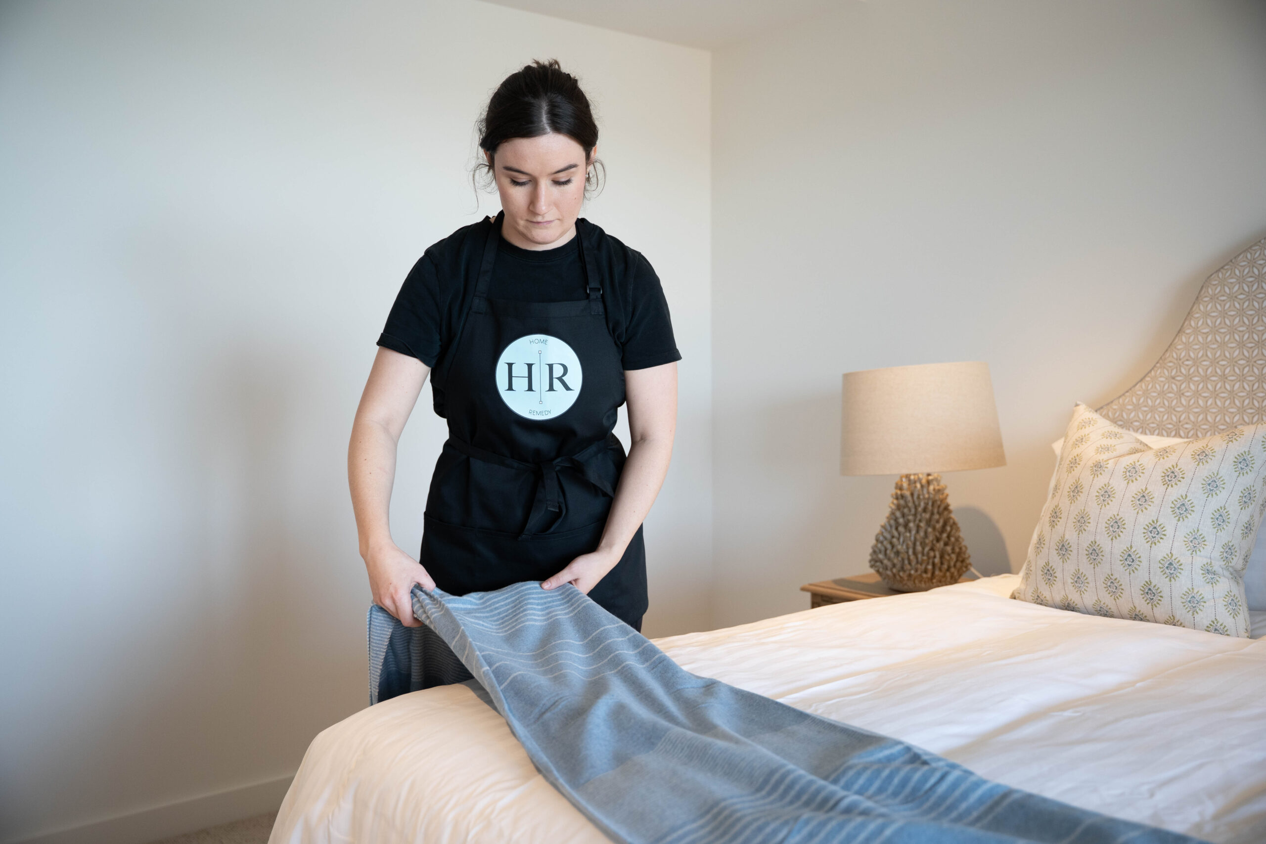 A woman from the Home Remedy team, wearing a black apron, is neatly arranging a gray throw blanket on a made bed in a bright, tidy bedroom.