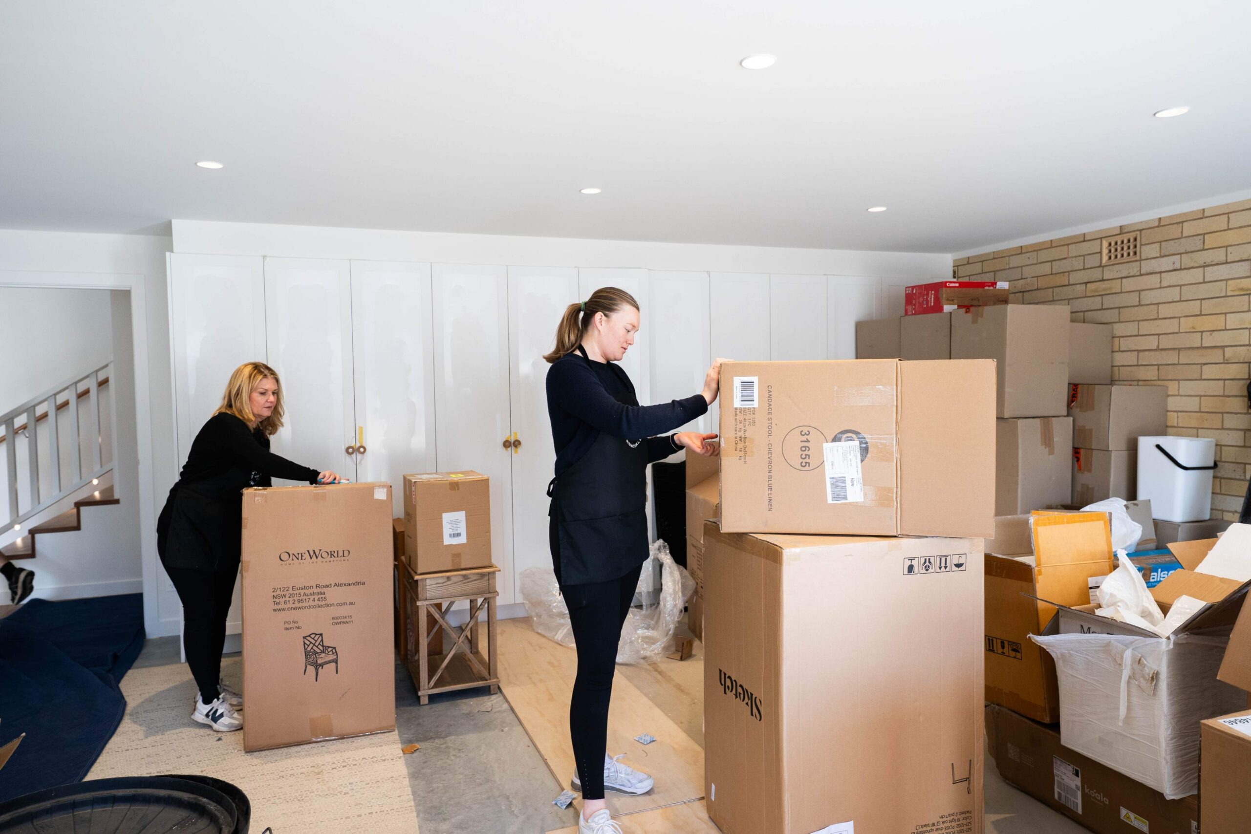 Two women stand in a room filled with large cardboard boxes, unpacking and organizing items in a brightly lit space with white cabinets in the background.
